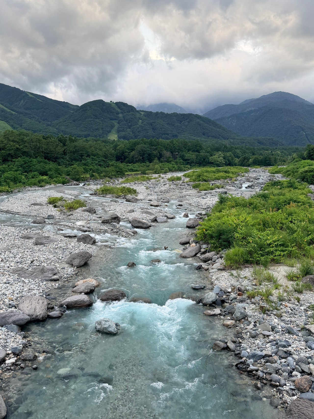 A pale blue river crosses a rocky bed beneath cloud-covered mountains