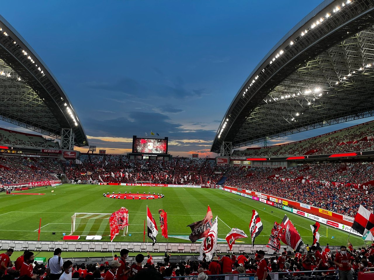 A football stadium at dusk with lit grandstands and spectators in red