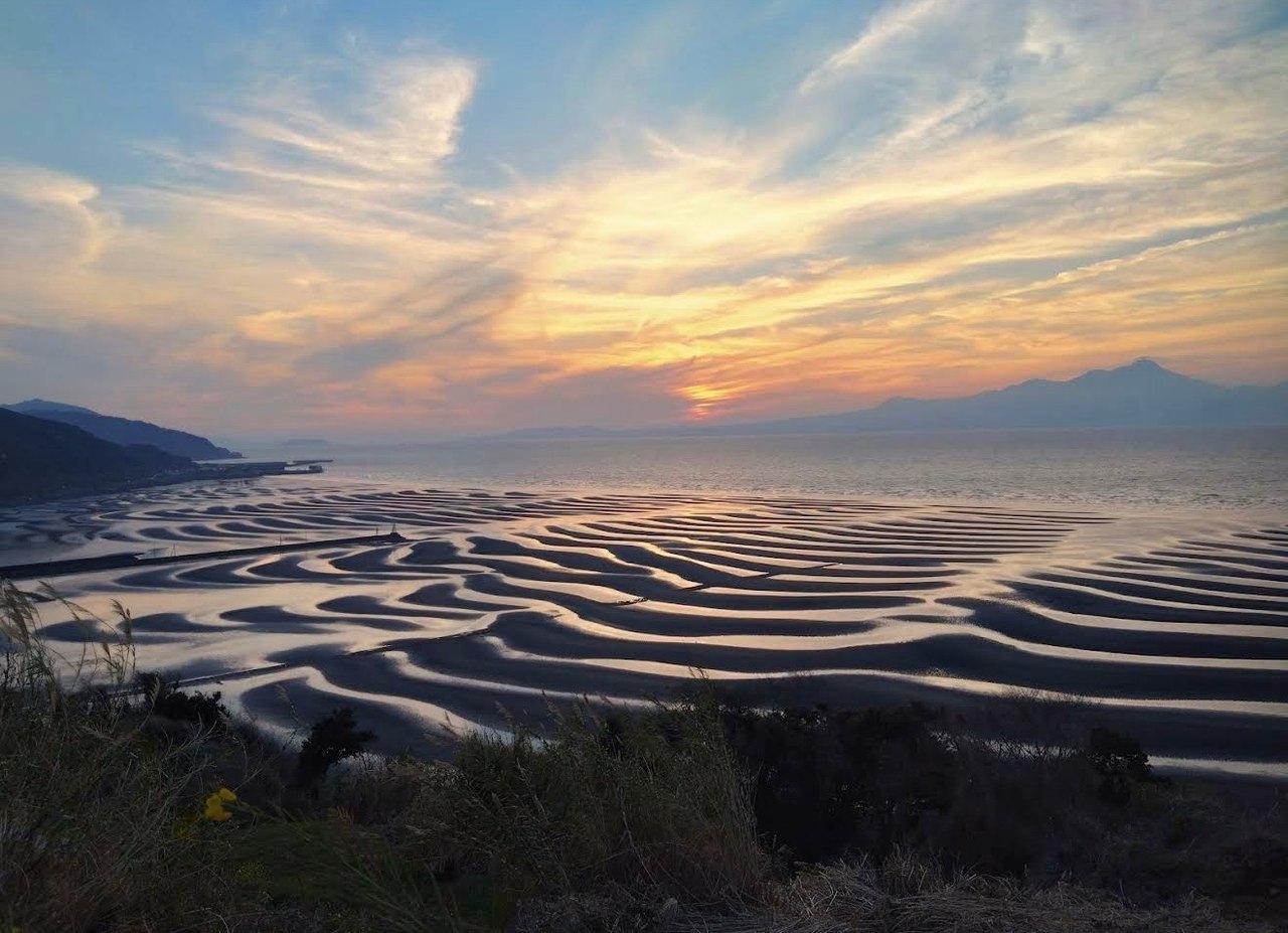 Curving bands of water cross tidal flats at sunset with a mountain beyond